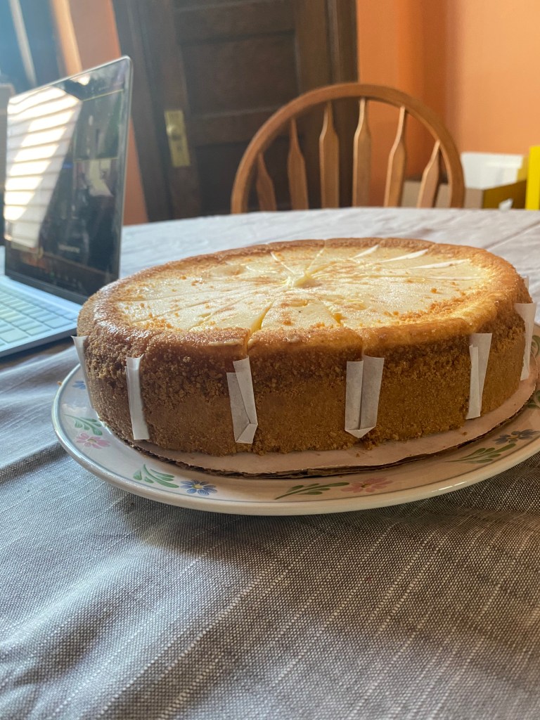 An enormous and delicious New York style cheesecake with a (gluten free) cookie crust sits atop a table covered in a grey tablecloth.